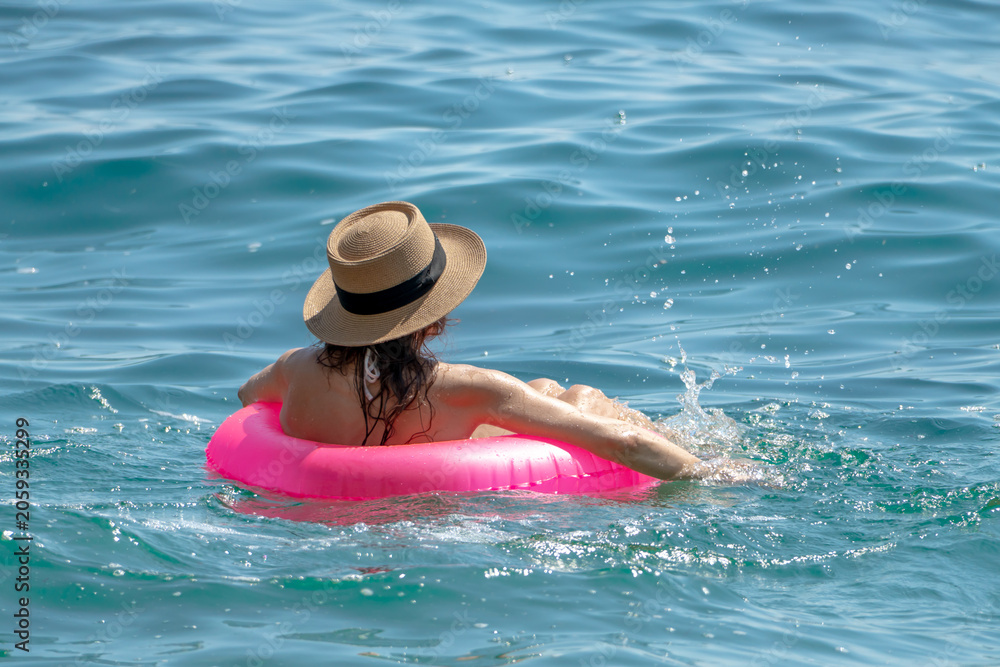 Naklejka premium Woman Swimming Sea: Woman in hat relaxing, floating pink tube, splashing warm ocean water on sunny day.