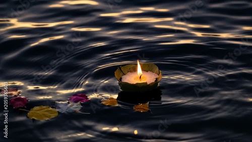 Gentle hand holds lit candle on calm dark water. Floating petals drift with the candle's glow. Soft golden light reflects on rippling surface. Peaceful ritual captures quiet reflection