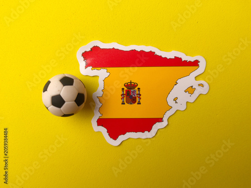 A Spain map sticker printed with its national flag design sits next to a miniature football on a yellow background