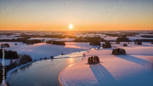 Aerial drone footage showing vast beautiful winter landscape at sunrise with winding frozen river reflecting golden light surrounded by snow covered fields and pine forests
