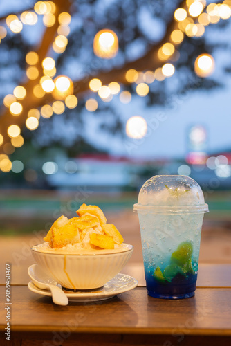 Фотография Delicious milk bingsu shaved ice topped with crispy butter toast cubes served with a blue Italian soda cocktail on a wooden table with gorgeous night bokeh lights