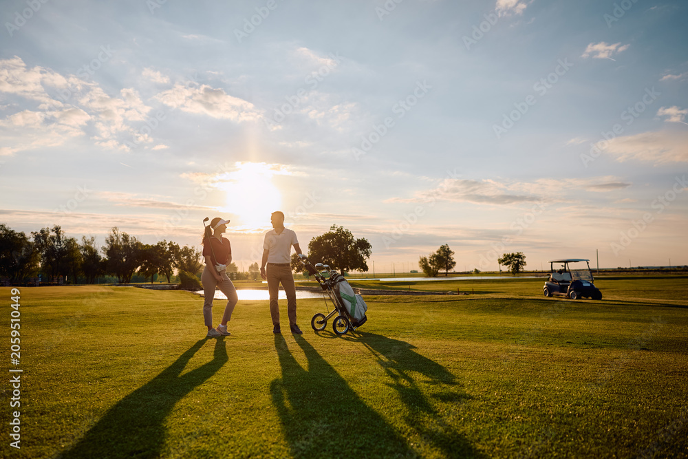 Naklejka premium Full length of happy golfers communicating on golf course at sunset.