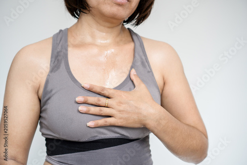 Sweaty Asian woman in grey sportswear holding her chest in pain after a workout at home, suffering from sudden chest tight, heart attack, breathing difficulty, or overtraining syndrome