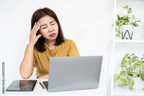 Stressed Asian woman touching her temple, suffering from an intense headache, migraine, or eyestrain while working with a laptop at her desk at home, feeling exhausted, tired, and burned out