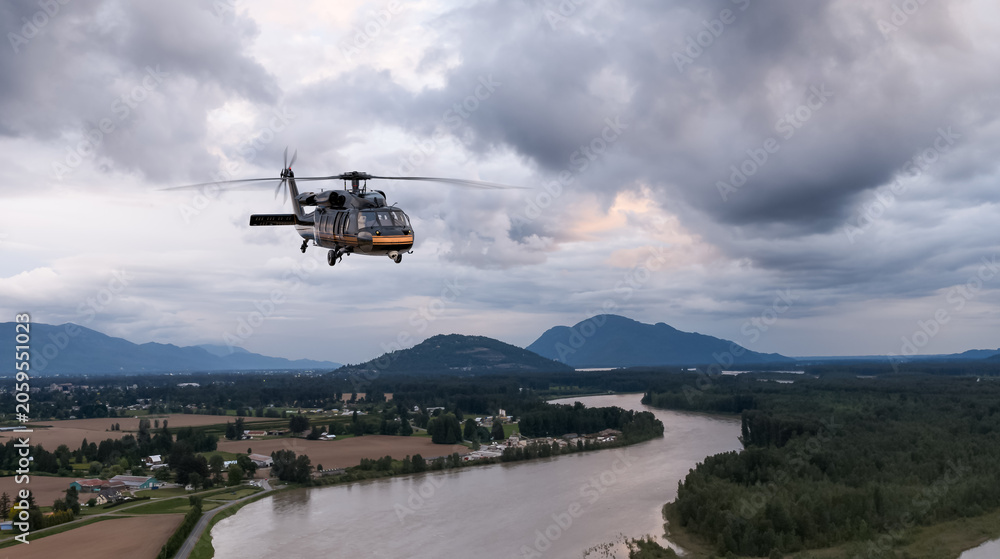 Fototapeta premium Military Helicopter Flying Over River Valley With Storm Clouds and Mountain Background in BC, Canada