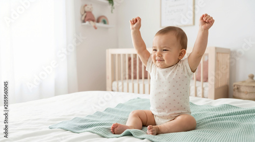 Happy baby stretching arms up while sitting on a bed in a bright nursery. Waking up in the morning on a green blanket with a wooden crib in the background