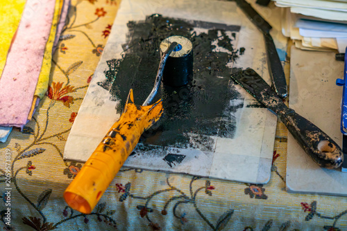 Printmaking workshop still life with a linocut roller featuring a dirty yellow handle, placed on an inking plate covered with black ink next to metal palette knives and spatulas on a patterned tablecl