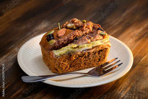 Homemade banana bread loaf topped with almonds, walnuts, and pumpkin seeds served on a white ceramic plate with a fork on wooden table. Delicious bakery concept, close up view.