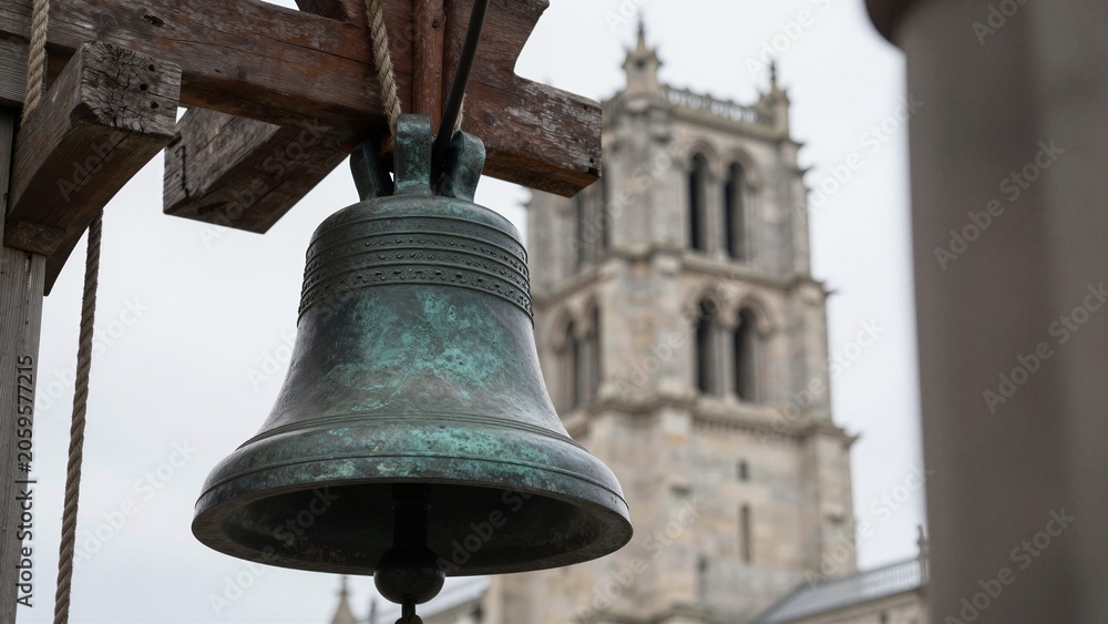 Obraz premium Close-up of an aged bronze church bell on a wooden frame, with a historic stone cathedral tower in the soft focus background.