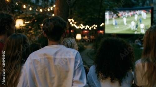 Group of friends watching soccer match on outdoor screen at night celebration