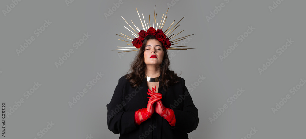 Obraz premium Photo banner. Young adult white woman with closed eyes, red lips, tulle gloves, make up, DIY golden halo crown, rose on head, choker, collar on neck, black jacket on gray. Beauty, pray, faith concept