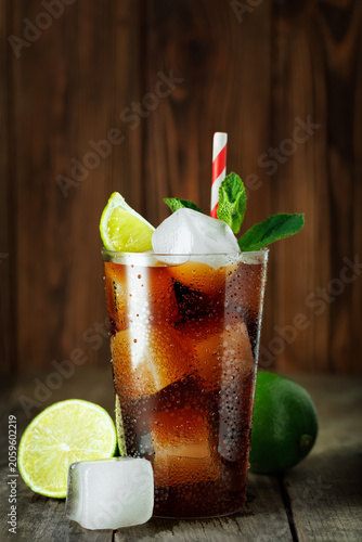 Cuba libre cocktail with rum, cola, fresh lime and mint, refreshing alcoholic drink served in glass with ice and striped straw on wooden background