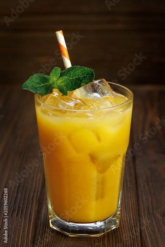 Orange juice in glass with ice cubes, fresh mint sprigs and striped straw on dark wooden table