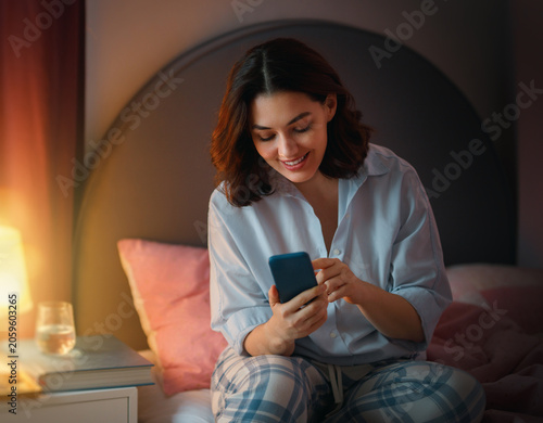 Happy Young Woman in Pajamas Using Smartphone in Cozy Bedroom