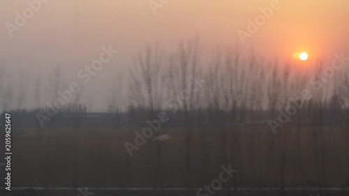 Looking out from train window, sun disk stuck in foggy air, naked trees flick by on foreground, Chinese farm fields seen at countryside. Ride at high speed train from Shanghai to Beijing
