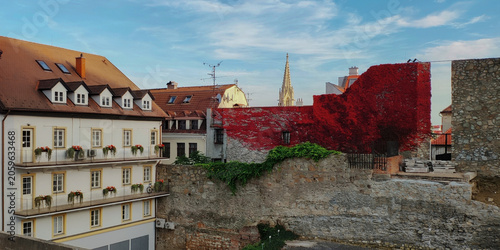 Urban scene with traditional buildings, striking red modern art installation on a wall and stone ruins under blue sky. 