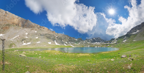 Beautiful mountain lake panorama under bright sunshine and blue cloudy sky. Scenic alpine landscape.