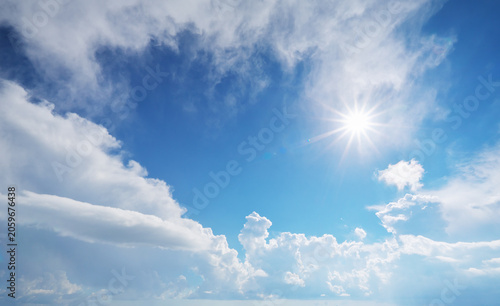 Bright sun burst with starburst rays shining through white fluffy clouds in blue sky. Beautiful sunny daytime sky background for commercial video production, religious or nature design template.
