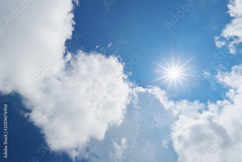 Bright sun burst with starburst rays shining through white fluffy clouds in blue sky.