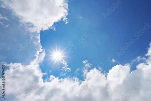 Bright sun burst with starburst rays shining through white fluffy clouds in blue sky.