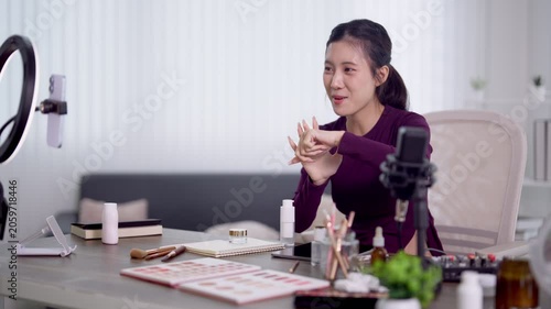 Young Asian woman podcaster presenting skincare product to camera while recording beauty podcast episode at home desk with ring light and microphone