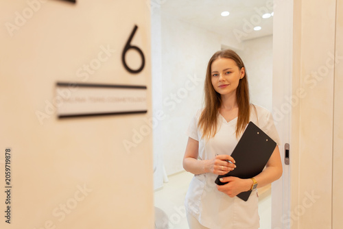 A young professional female esthetician or dermatologist in white scrubs holding a clipboard, standing in the doorway of a modern cosmetology clinic or medical office room number 6.