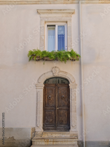 Charming Italian building facade showing a vintage wood doorway beneath a window with green planters. Picturesque travel photography from the historic streets of Ortigia, Sicily.