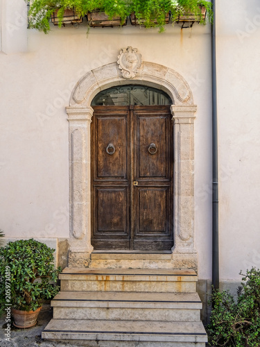Classic arched wooden door with stone portal frame and steps in Ortigia, Syracuse, Sicily, Italy. Historic Mediterranean building entrance with decorative details and potted plants.