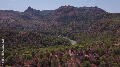 Drone footage descending toward El Chorro's jagged peaks, pulling the rugged limestone ridgeline and thick woodland canopy into sharp focus under intense summer light.