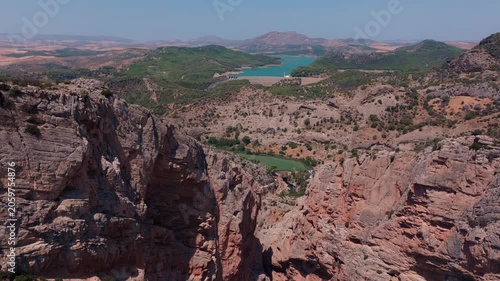 Drone footage drifting sideways and tilting down past jagged limestone pinnacles, opening to a vast valley with turquoise reservoir, rolling hills, and hazy mountain horizon beyond.