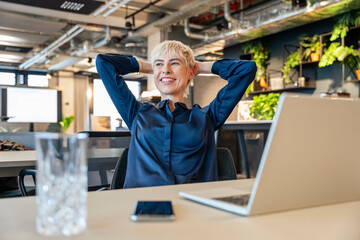 Smiling businesswoman relaxing at desk with laptop in modern office