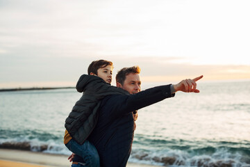 Father carrying son piggyback on the beach at sunset pointing finger