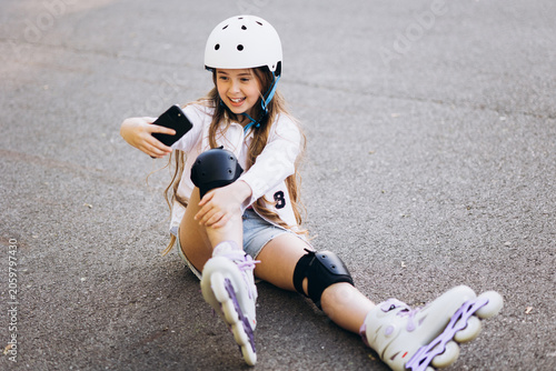 Happy girl wearing helmet and safety pads, sitting on asphalt road while taking a picture with smartphone