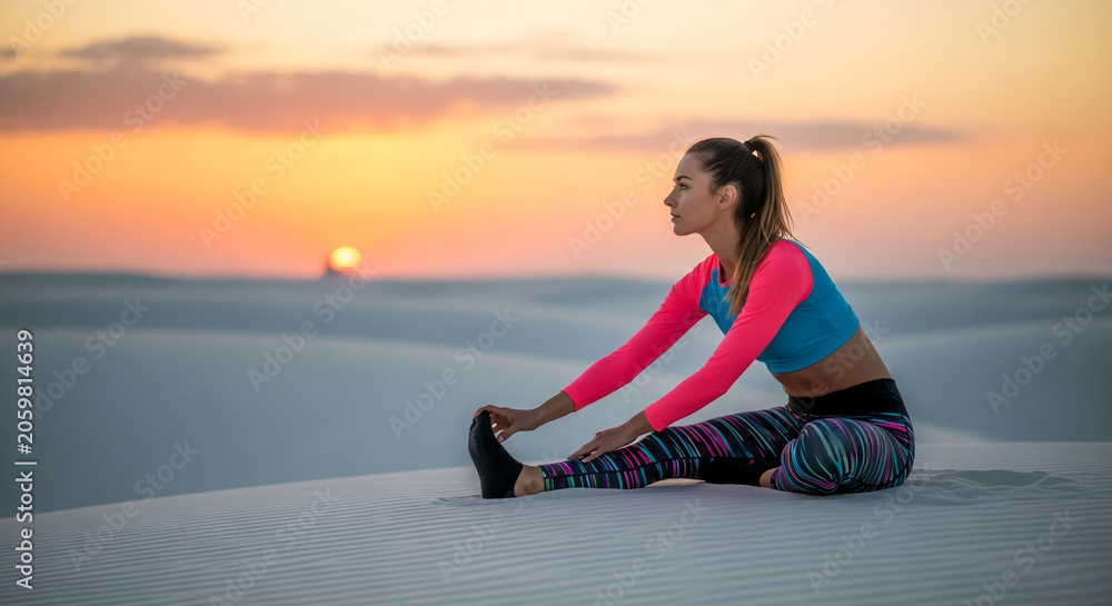 Fototapeta premium Woman stretching in colorful athletic wear on sandy dunes at sunset, symbolizing fitness, freedom, and outdoor activity.