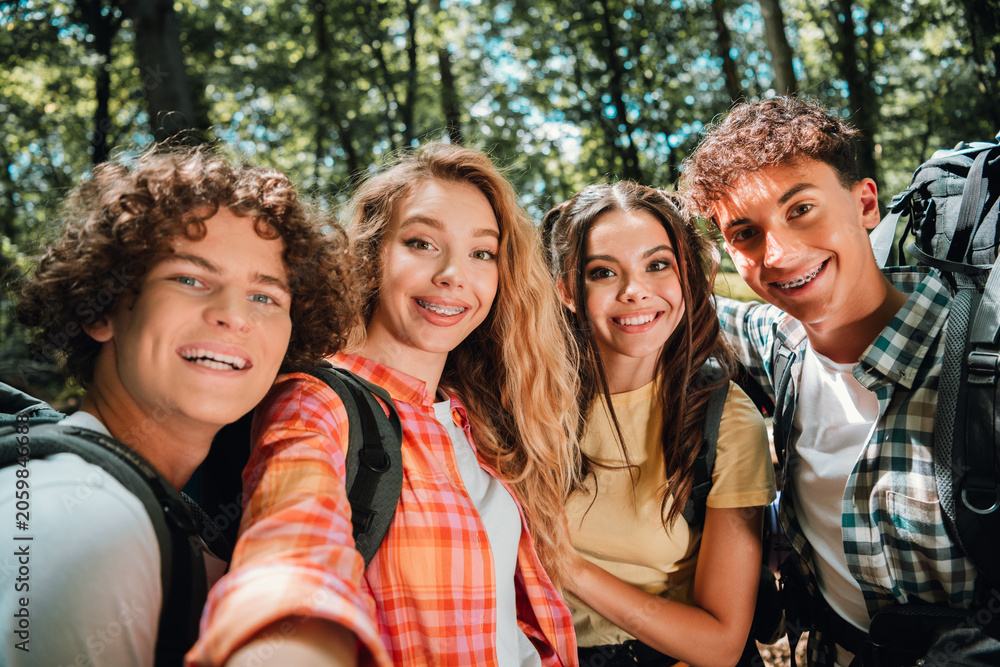 Obraz premium Four smiling teenage friends taking a group selfie while hiking in a sunny forest with backpacks and casual summer clothes
