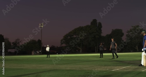 Asian man running and bowling ball testing teenage batsman on pitch, floodlights, copy space