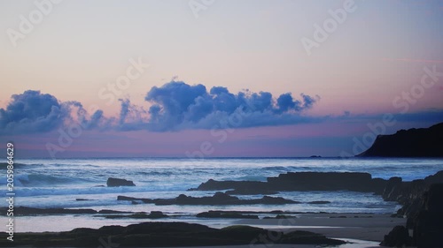 Beautiful 4K slow motion footage of the ocean at twilight with dramatic clouds moving over a rocky coastline. The peaceful scene captures the serene afterglow and the gentle movement of the waves agai