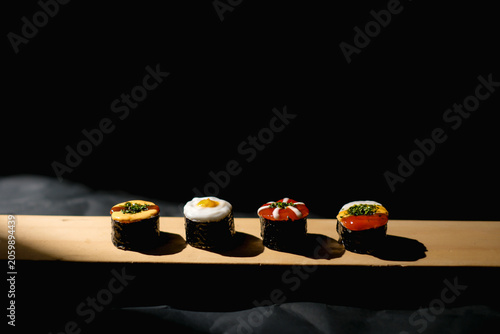 Row of four assorted creative Japanese fusion sushi rolls with various colorful toppings served on a long wooden board against a dark dramatic background.