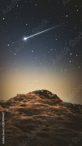 A breathtaking view of a bright shooting star or meteor streaking across a clear, starry night sky above a dark, textured rocky hill. Beautiful cosmic landscape