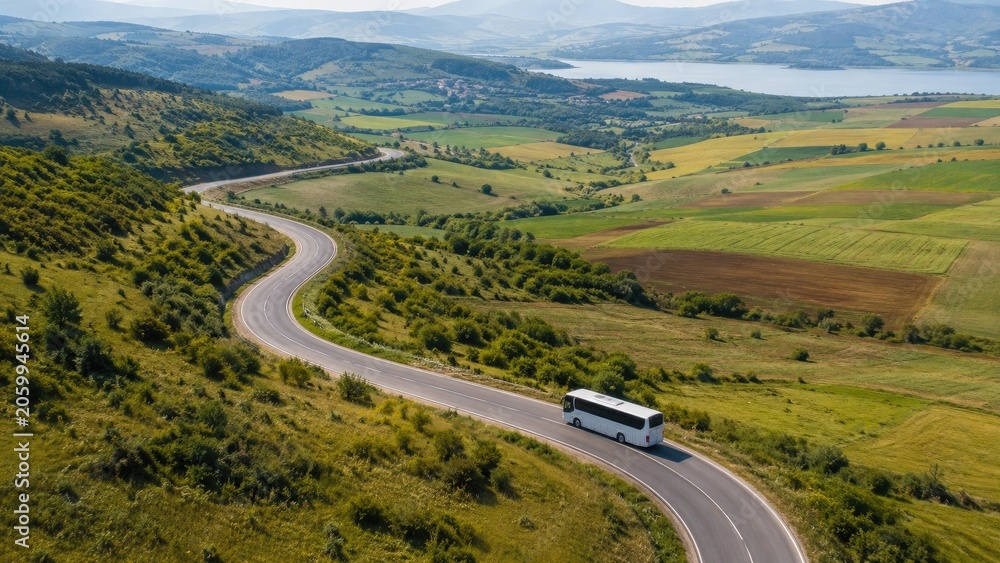 Fototapeta premium A white coach bus drives along a winding road through green hills, farmland and a lakeside valley, captured in an aerial panoramic landscape view