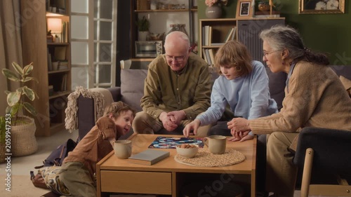 Long shot of senior Caucasian grandparents playing board game with young grandsons in living room. Clip showcases multigenerational family bonding, perfect for retirement planning or toy commercials