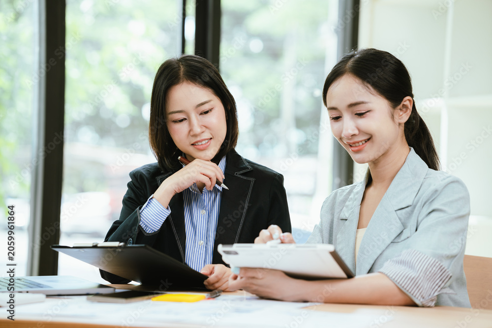 Obraz premium Two businesswomen collaborating at a modern office desk, discussing a project while reviewing information on a laptop and tablet in a bright professional workspace.