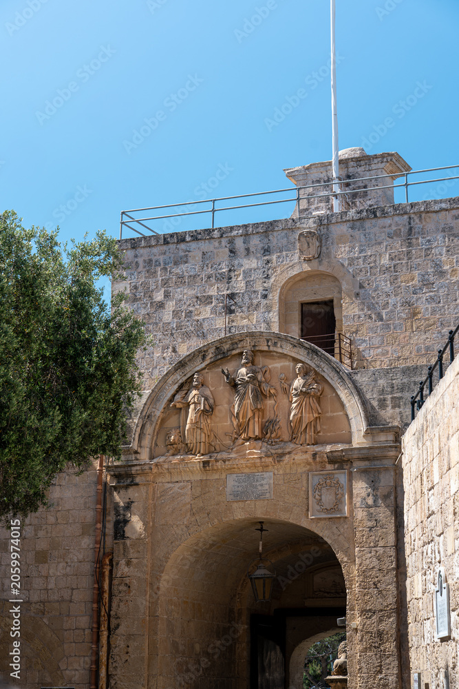Obraz premium view from behind mdina gate, malta with limestone brick and detailed statues above the archway with clear blue skies overhead in summer sunlight and green lush tree to the left
