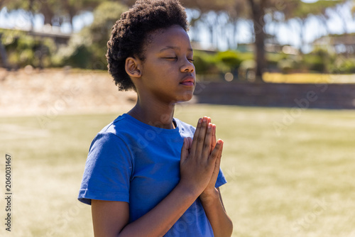 African American child standing on grassy park field in blue T-shirt pressing hands at chest