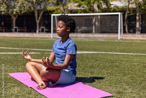 African American teen in blue sitting meditating on pink yoga mat on turf near white goal