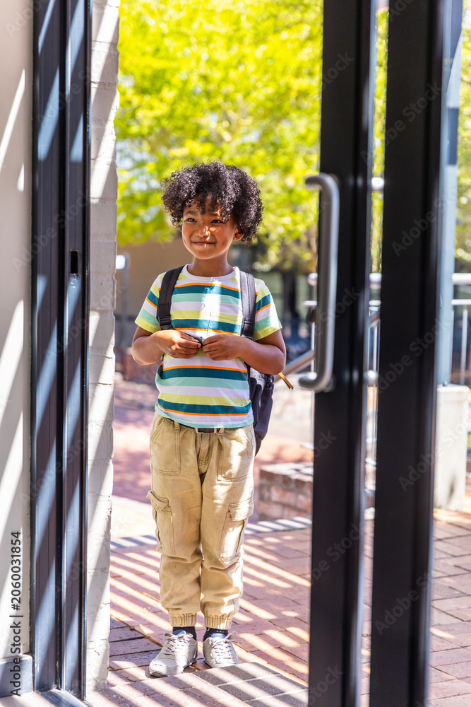 Obraz premium African American child standing in glass doorway carrying dark backpack and wearing striped T-shirt
