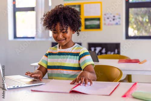 African American boy child sitting at school desk wearing striped Tshirt using laptop and notebook
