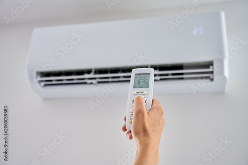 Hand changes air conditioner temperature with a remote control, setting the wall unit to 23 degrees. Concept of room cooling and indoor comfort.