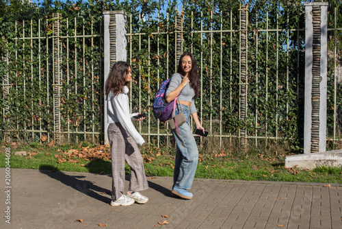 Mother with teenager walk together along a path beside a green fence in a city during a sunny day in autumn