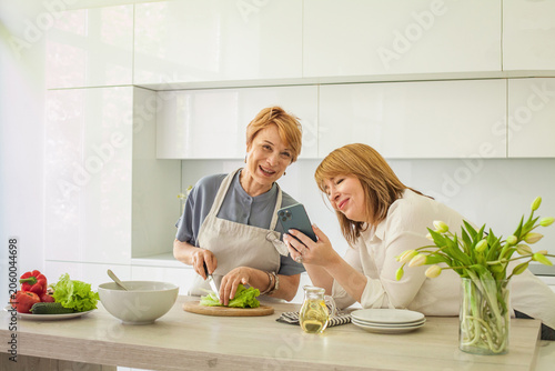 Two women cooking in a modern kitchen one slicing lettuce while the other checks a phone for a recipe in bright sunlight